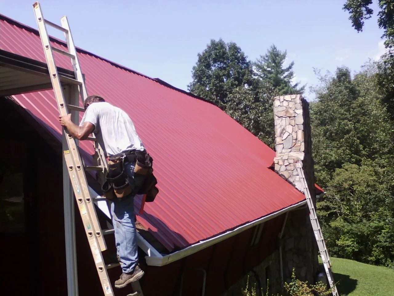 Roofer on a ladder beside a red metal roof and stone chimney — Soffit & Fascia Repair by Roofing Brothers in Clarkton